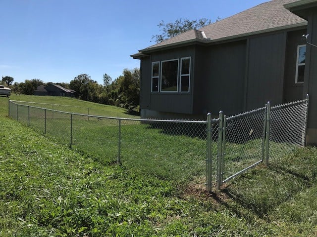 A fenced backyard with green grass beside a gray house under clear blue sky.